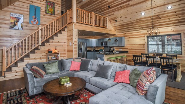 A spacious living area with a gray sectional sofa, a wooden staircase, and a kitchen in the background. - Woods-stock by Taylor Made Deep Creek (McHenry)