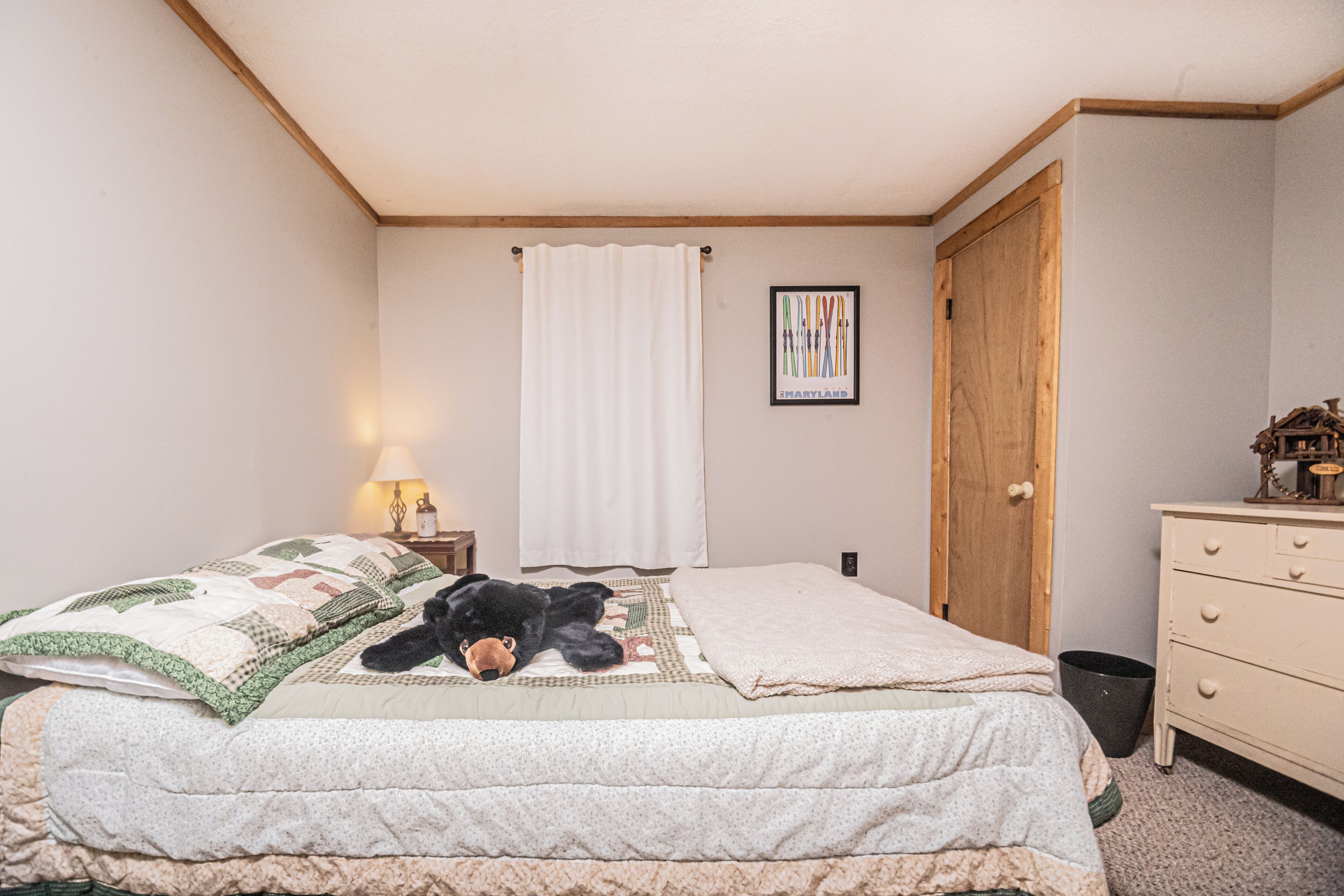 A bedroom with a bed, a teddy bear, a nightstand with a lamp, a wooden door, and a framed picture on the wall.