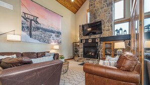 A cozy living room with leather sofas, a stone fireplace, a large painting, and a television mounted on the wall. - Bet Your Ace by Taylor Made Deep Creek (McHenry)