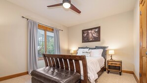 A bedroom with a bed, a wooden bench, a ceiling fan, a window with curtains, a bedside table with a lamp, and a framed picture on the wall. - Bet Your Ace by Taylor Made Deep Creek (McHenry)
