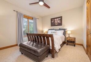 A bedroom with a bed, a wooden bench, a ceiling fan, a window with curtains, a bedside table with a lamp, and a framed picture on the wall. - Bet Your Ace by Taylor Made Deep Creek (McHenry)