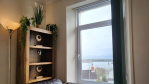 A living room with a leather sofa, a wooden bookshelf with plants, and a view of a bridge over water through a window. - Marina Reach, Neyland, Pembrokeshire: Sea views (Neyland)
