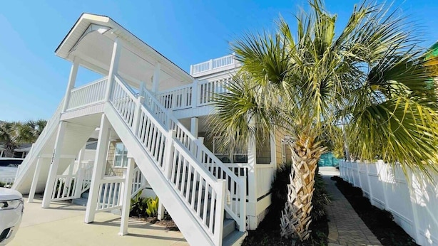 A two-story building with a white exterior, a staircase leading to the upper level, and a palm tree in the foreground. - Spacious Beach Duplex | 5 Bedrooms | Pool  #B (North Myrtle Beach)