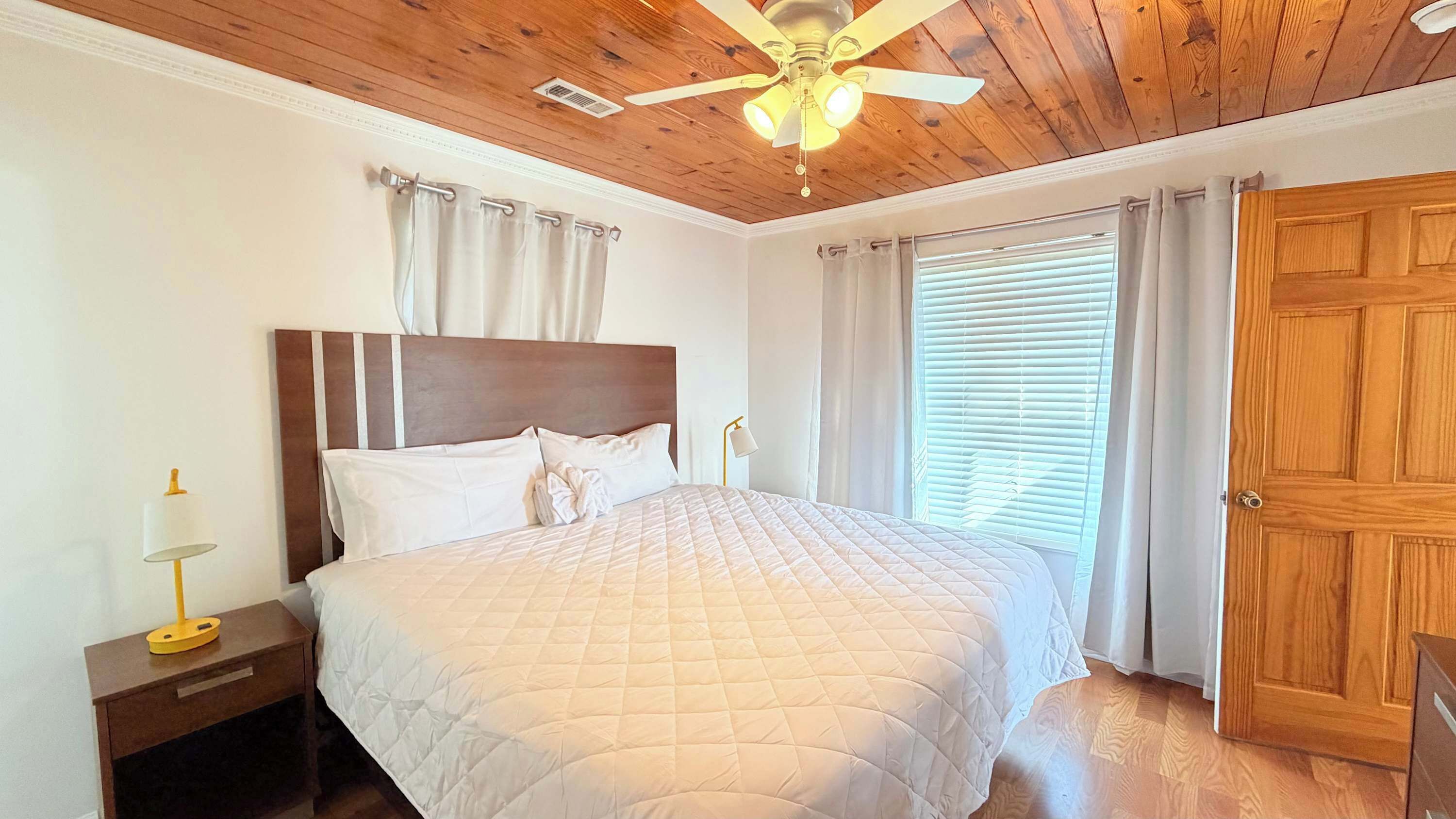 Une chambre avec un plafond en bois, un lit recouvert d’une couette matelassée, une table de chevet avec une lampe et une porte.