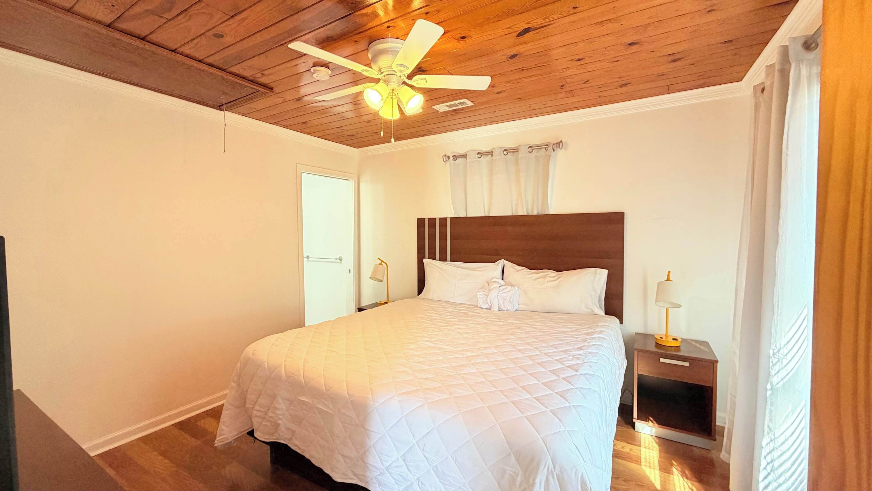 Une chambre avec un plafond en bois, un lit recouvert de draps blancs, une table de chevet avec une lampe et une fenêtre avec des rideaux.