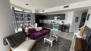 A modern living room with a purple sofa, a grey armchair, and a glass coffee table. A kitchen is visible in the background with dark cabinets and stainless steel appliances.