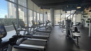 A gym with treadmills, exercise bikes, and weight machines. Large windows offer views of tall buildings.