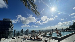 A rooftop pool area with lounge chairs and a swimming pool, surrounded by high-rise buildings and palm trees.