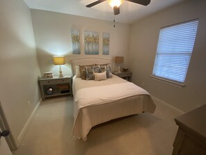 A bedroom with a bed, nightstands, a ceiling fan, artwork on the wall, and a window with blinds.