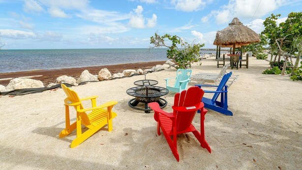 Uma praia com uma estrutura de telhado de palha, uma lareira e cadeiras Adirondack coloridas.
