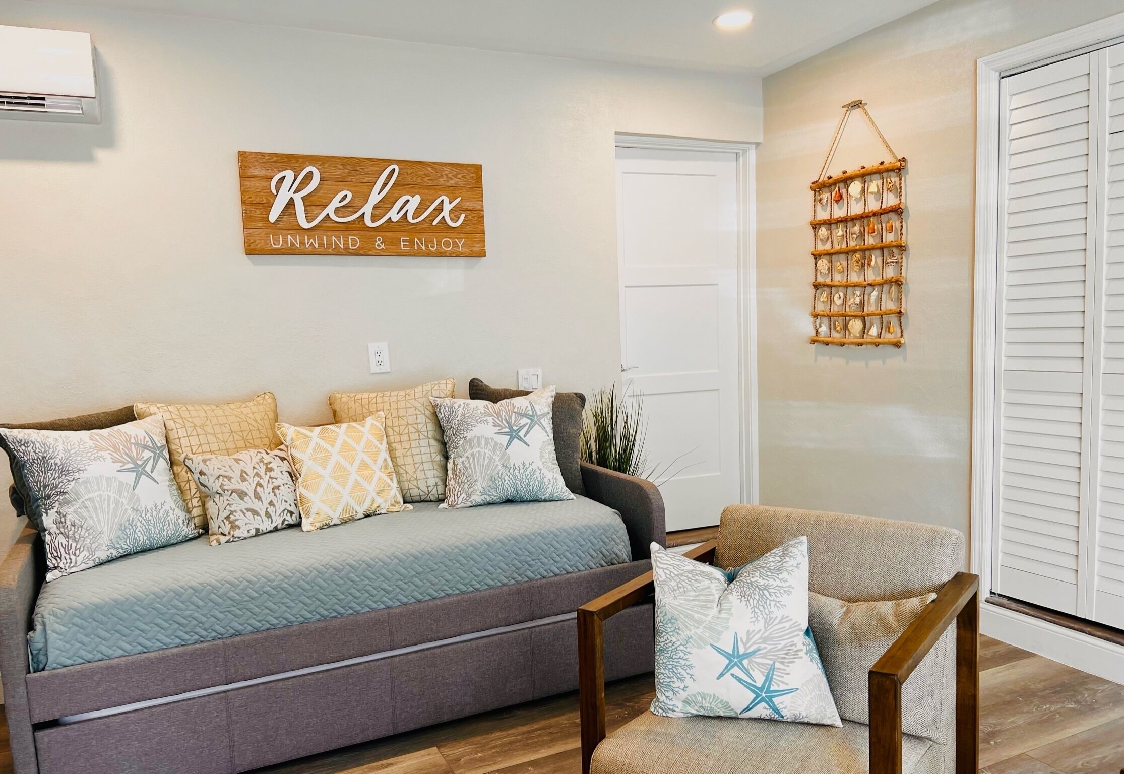 A living room with a sofa and armchair, a wall-mounted sign that reads "Relax Unwind & Enjoy," and a decorative wall hanging.