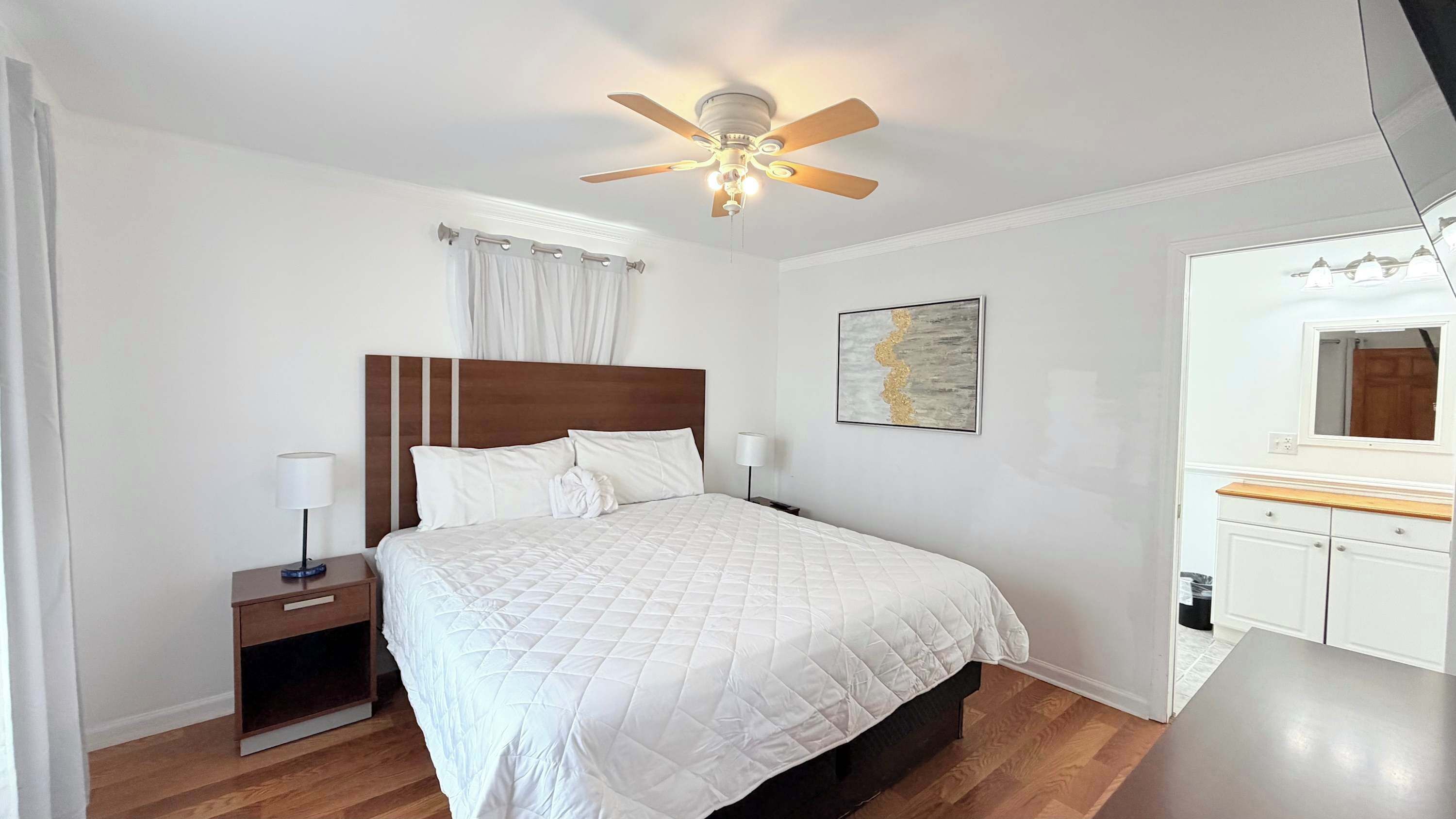 A bedroom with a bed, bedside tables, a ceiling fan, a nightstand, a wall-mounted lamp, a painting, and a mirror.