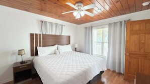 Une chambre avec un grand lit, un plafond en bois, un ventilateur de plafond, une table de chevet avec une lampe et une fenêtre avec des rideaux.