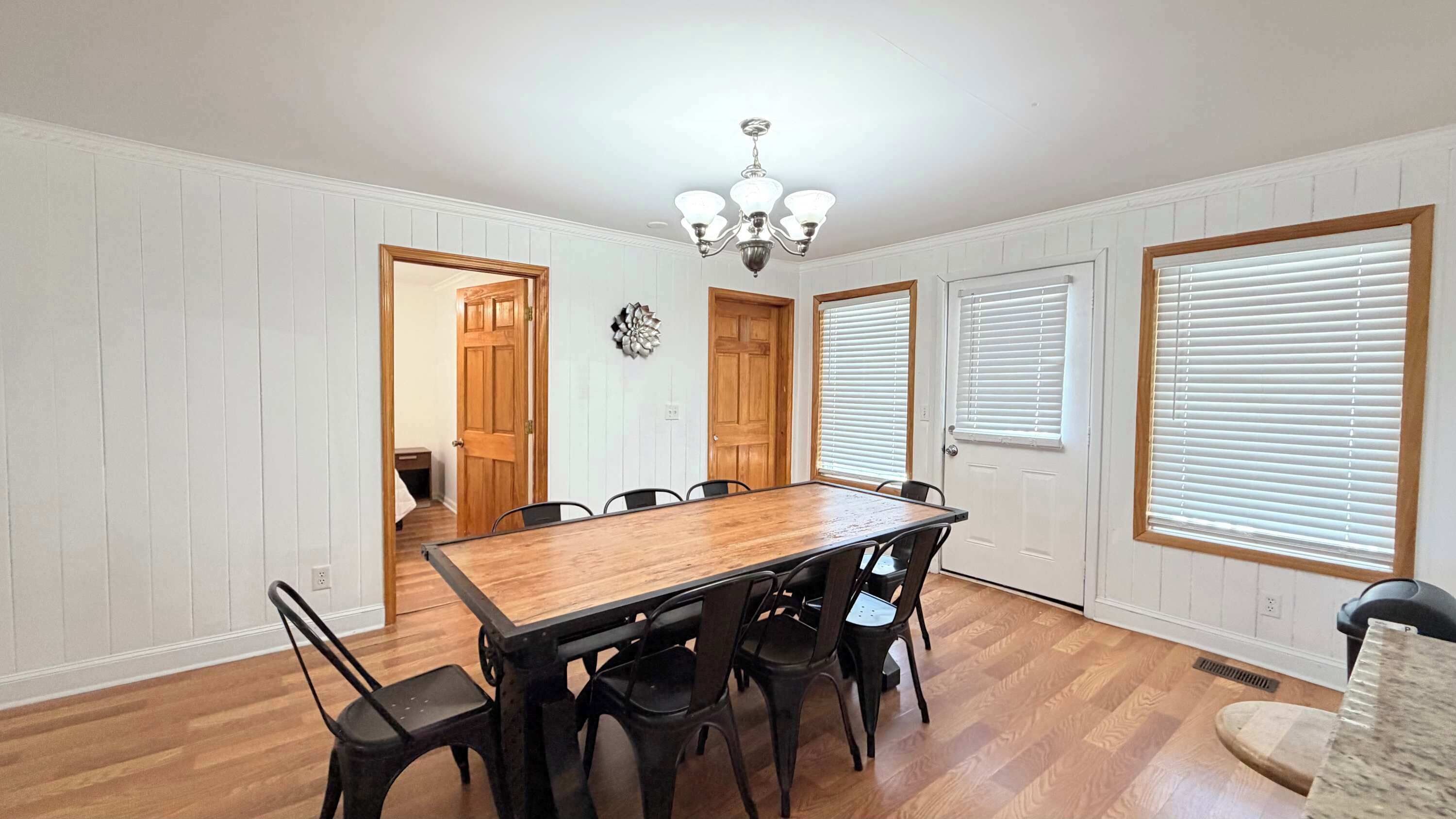 Comedor con mesa y sillas de madera, paredes blancas con paneles y una araña.