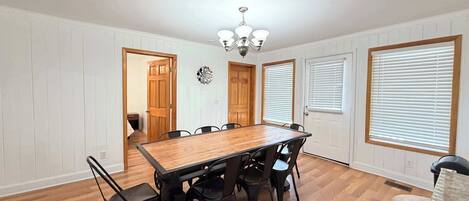 Comedor con mesa y sillas de madera, paredes blancas con paneles y una araña.