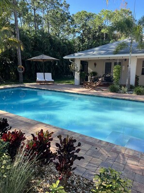 A swimming pool with a patio area, surrounded by lush greenery and a house with a covered patio.