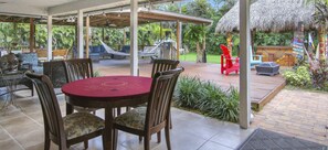 A covered outdoor area with a wooden deck, a round table with chairs, and a thatched-roof structure in the background.
