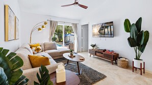 A living room with a sofa, coffee table, and a ceiling fan. There is a TV mounted on the wall and a window with curtains. - *NEW 3/2 Pool Home, Private Yard, Great Location! (Jupiter)