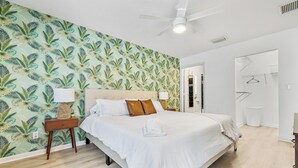 A neatly made bed with white linens, a wooden nightstand with a lamp, and a tropical leaf-patterned wall. - *NEW 3/2 Pool Home, Private Yard, Great Location! (Jupiter)