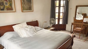 Een opgemaakt bed met houten hoofdbord, een nachtkastje met een lamp, een staande ventilator, een houten stoel en een spiegel aan de muur.