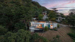 A modern house with multiple levels, surrounded by dense forest, featuring a large outdoor deck and a visible staircase.