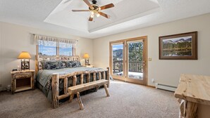 A bedroom with a wooden bed, two bedside lamps, a ceiling fan, and a large window with a view of the outdoors. - Lily Mountain Lodge - Amazing Mountains Views, Private Hot Tub, Great Location (Estes Park)