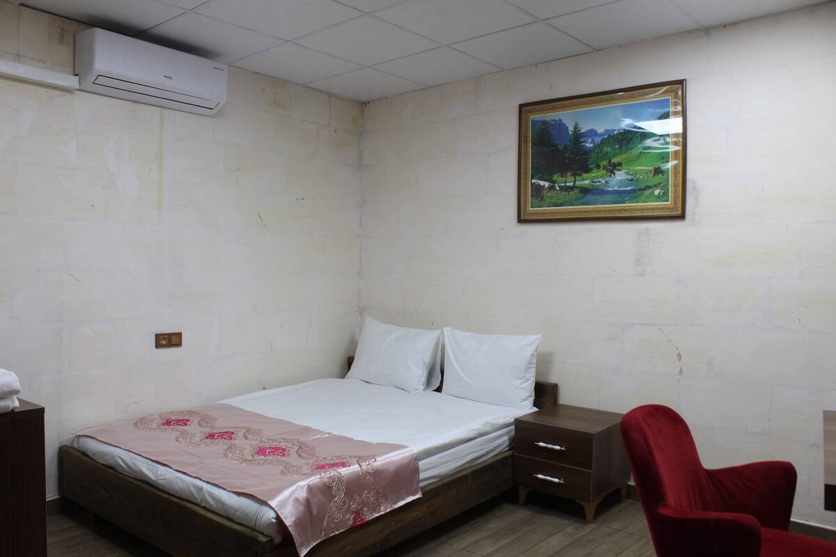 A bedroom with a bed, a nightstand, a red chair, an air conditioner, and a framed painting on the wall.