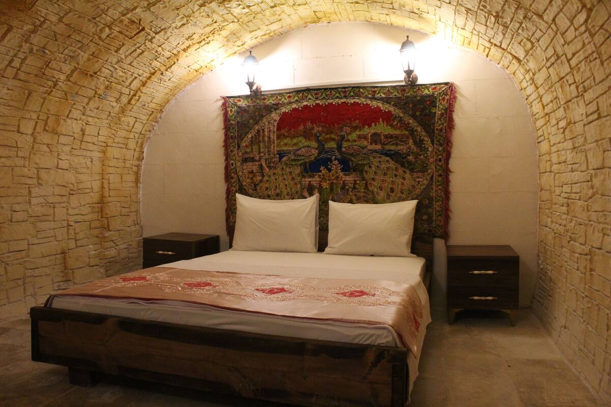 A bedroom with a stone arched ceiling, a large tapestry on the wall, a bed with white linens, and two bedside tables.