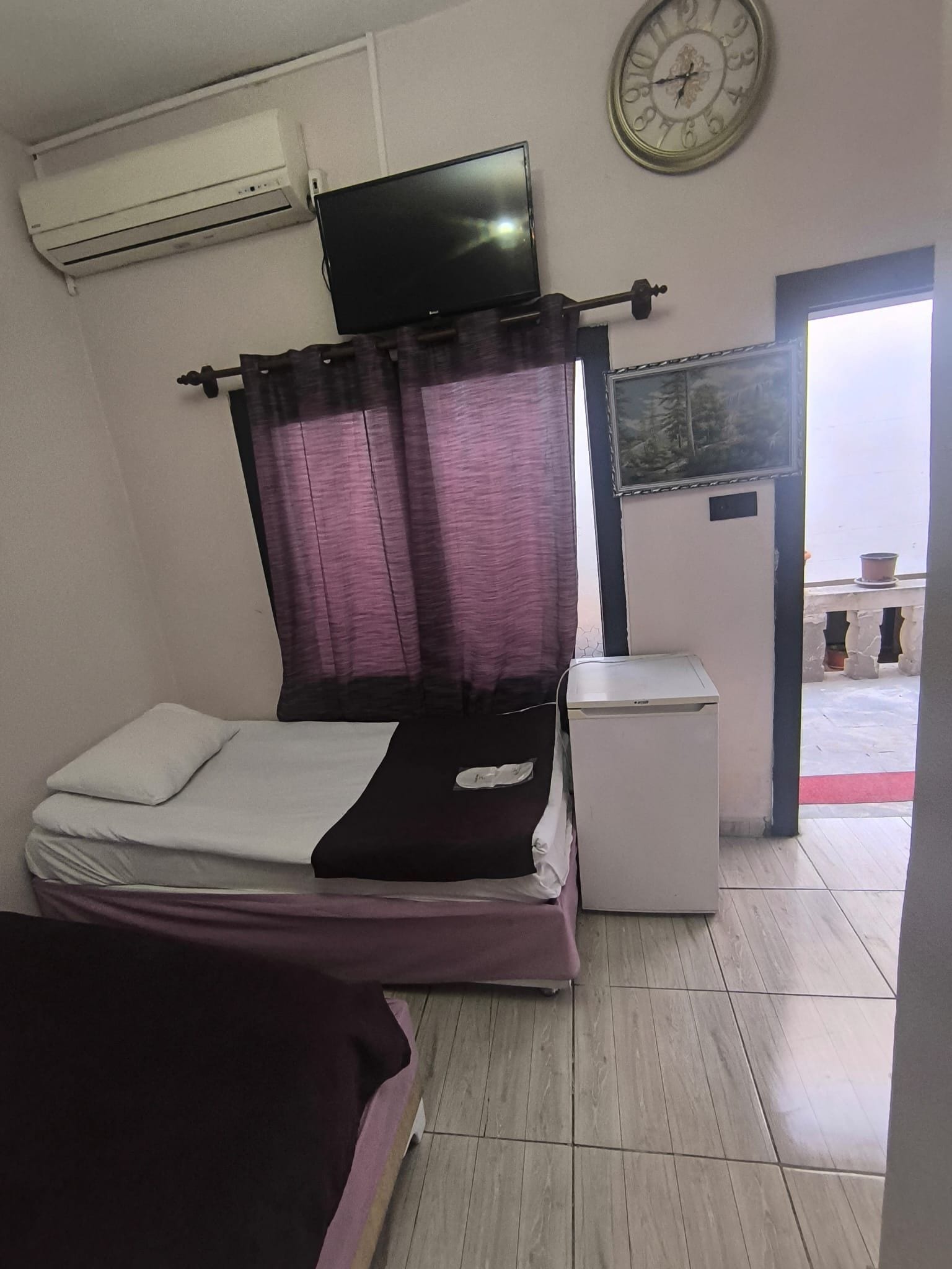 Quarto com cama, televisão presa na parede, mesinha de cabeceira e janela com cortinas.