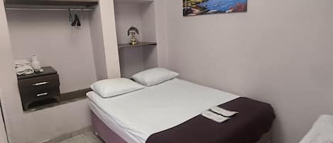 A bedroom with a bed, a nightstand, a shelf with decorative items, a painting, and a patterned rug.