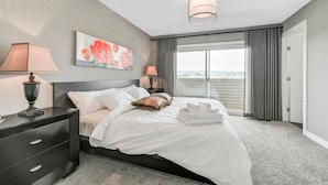 A hotel room with a bed, two bedside lamps, a painting, a large window with curtains, and a view of the outside.