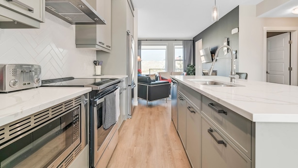 A modern kitchen with stainless steel appliances, a herringbone tile backsplash, and a view to the living area.