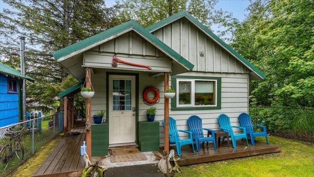 Eine kleine Holzhütte mit einer Veranda, einer Tür und einem Fenster. Auf der Veranda stehen vier blaue Stühle.