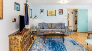 Ein Wohnzimmer mit Sofa, Couchtisch und Fernseher. An den Wänden hängen Bilder in Rahmen, und es gibt ein Holzregal.