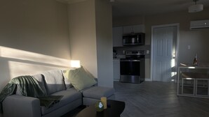 Un salón moderno con un sofá gris, una mesa de centro y, al fondo, una cocina con microondas y horno.