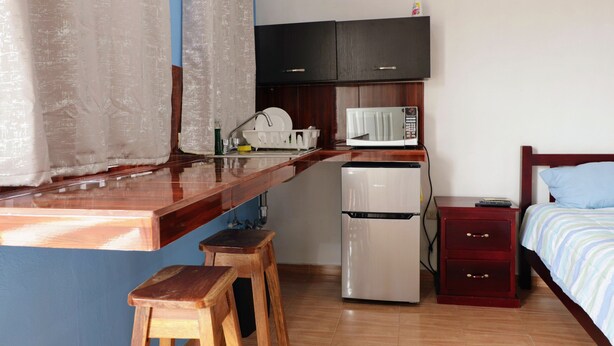 A compact kitchen with a refrigerator, microwave, and a small countertop. There is a wooden stool and a bed with a striped blanket. - San Román room inside the historic center (Campeche)