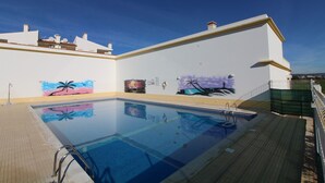 A swimming pool with a tiled deck, a white building with murals, and a clear blue sky. - Falésia Beach - Wifi, Piscina e Varanda (Albufeira)