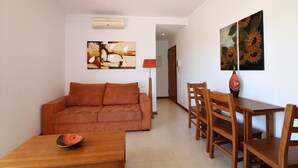 A living room with a sofa, a dining table with chairs, and two wall-mounted paintings. - Falésia Beach - Wifi, Piscina e Varanda (Albufeira)