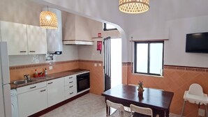 Une cuisine de taille réduite avec des meubles blancs, une cuisinière et un évier. On y trouve une table à manger avec des chaises et un téléviseur fixé au mur.