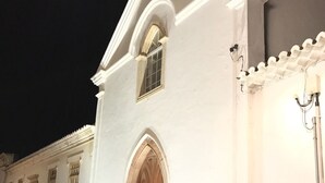 Un bâtiment blanc avec une grande porte en bois et des fenêtres en arc de cercle, illuminé la nuit.