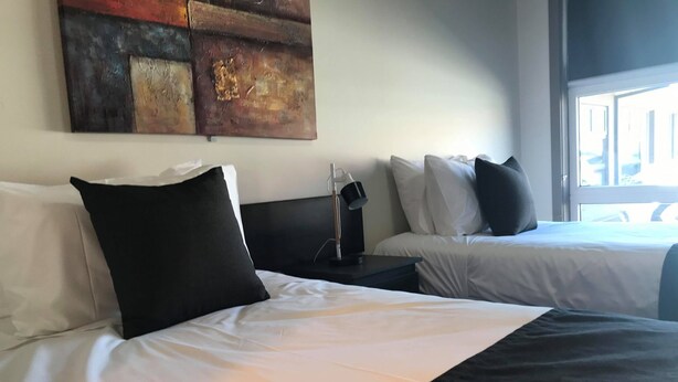 Habitación de hotel con dos camas, cada una con ropa blanca y almohadas negras. Hay un cuadro en la pared sobre las camas y una ventana que proporciona luz natural.