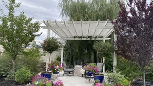 Uma área de varanda coberta com móveis de exterior, rodeada por um jardim com diversas plantas e flores.