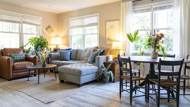 Een woonkamer met een bruine leren bank, een grijze hoekbank en een eettafel met stoelen. Er staan potplanten en een vloerkleed op de vloer.