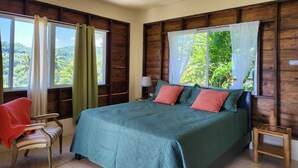 Une chambre avec un grand lit, une chaise et une petite table. Les fenêtres sont dotées de rideaux et offrent une vue sur la verdure.