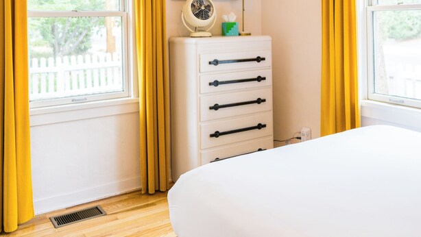 Ein Schlafzimmer mit Bett, Kommode, Ventilator und einem Fenster mit Vorhängen.