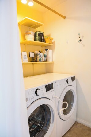 A laundry room with a front-loading washing machine and dryer, shelves above containing laundry supplies, and a wall-mounted light fixture.