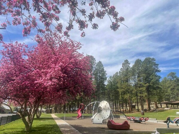 Un parque con un gran árbol de flores rosadas, una escultura de metal y un área de juegos.