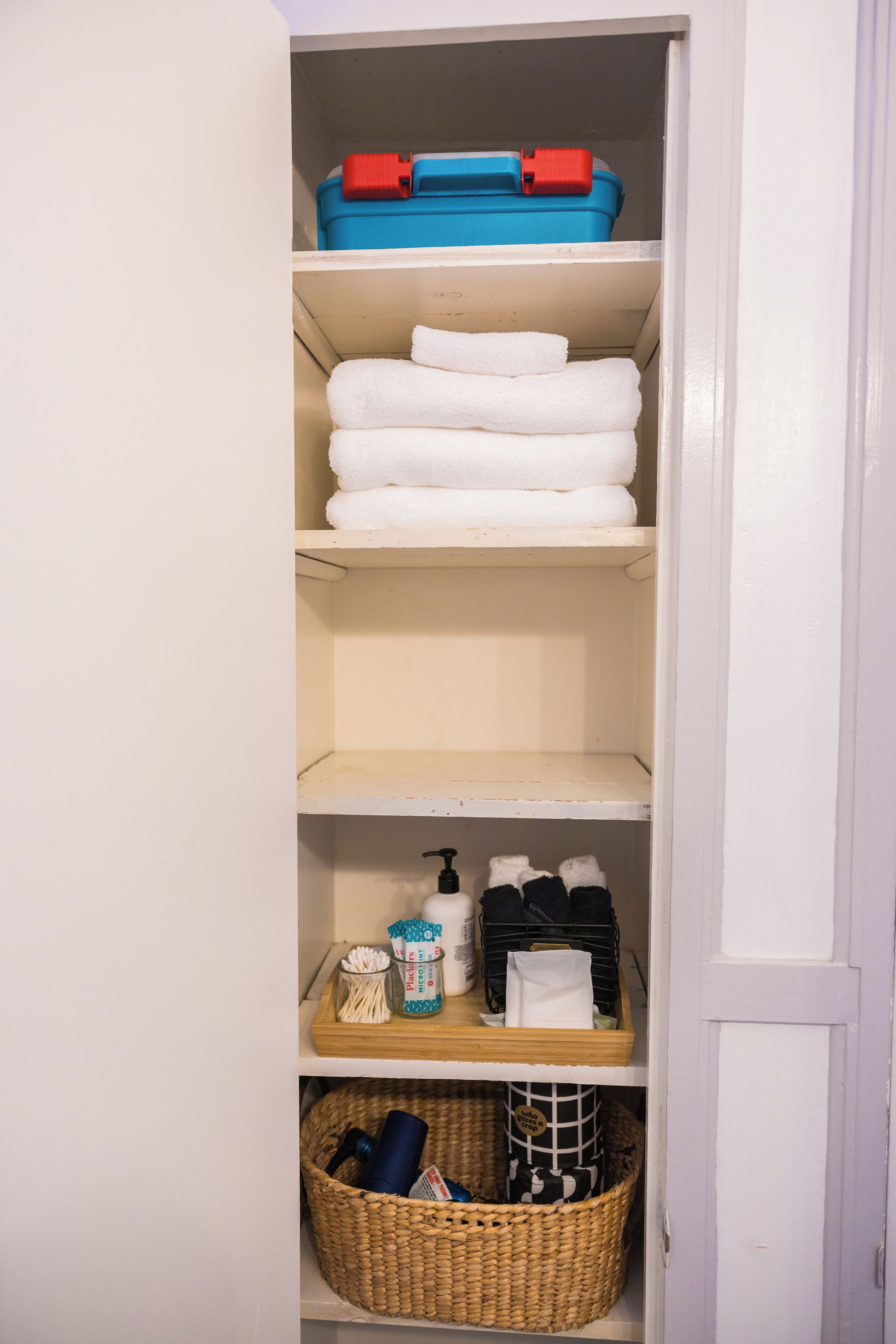 A storage closet with shelves containing folded white towels, a blue and red toiletry case, a basket with cleaning supplies, and a wicker basket with various items.