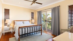 A bedroom with a bed, two bedside tables, a ceiling fan, a balcony with curtains, and a view of palm trees. - The Beachaus - Spacious Beachfront Holiday Home (Moreton Island)