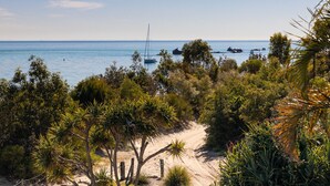 A coastal landscape with a sandy path leading to the sea, surrounded by dense vegetation and boats anchored in the water. - The Beachaus - Spacious Beachfront Holiday Home (Moreton Island)